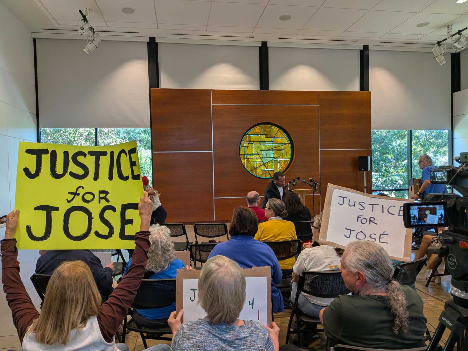 Crowd gathers in a modern meeting room holding bright signs reading 'JUSTICE FOR JOSE' as a man speaks at a podium in front of a circular stained-glass wall panel.
