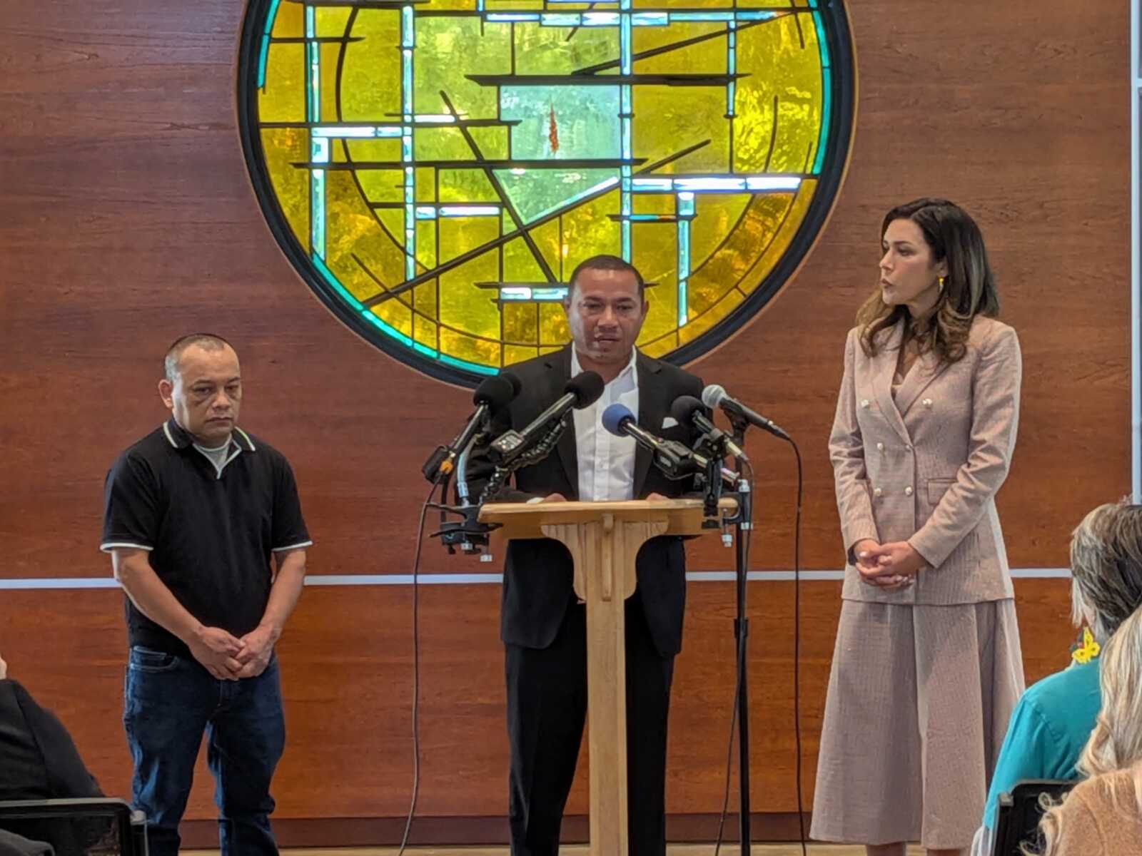 A man in a suit speaks at a wooden lectern with multiple microphones, flanked by a woman in a light pink suit, in a room with a large circular stained-glass window behind them.