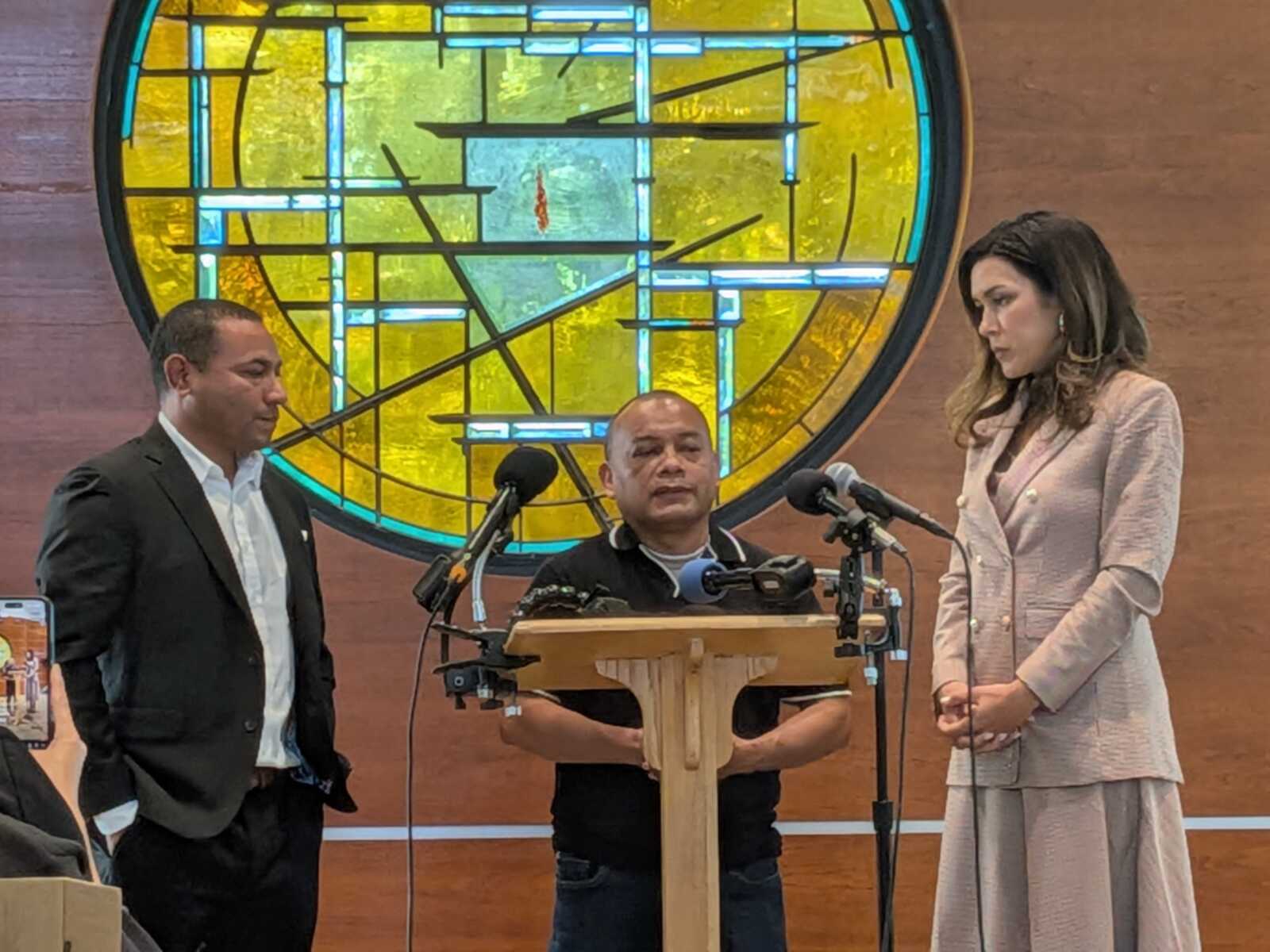 Three people at a press conference: a man at a wooden podium flanked by a man in a suit left and a woman in a light pink blazer right, with a colorful circular stained-glass window behind them.