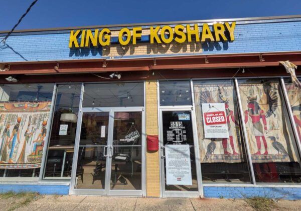 Front of a blue storefront reading 'KING OF KOSHARY' in large yellow letters above glass doors; Egyptian-themed banners in the windows.