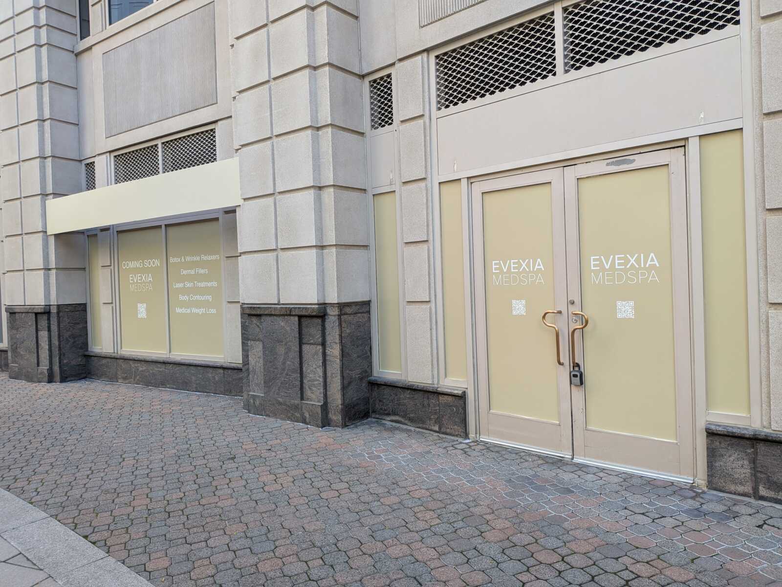 Modern storefront on a stone building with frosted glass doors reading EVEXIA MEDSPA; beige panels and gold handles, left window lists services.