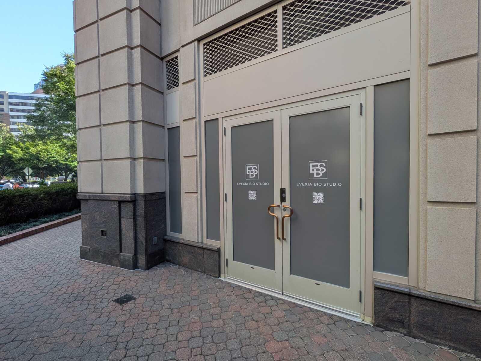 Entrance to Evexia Bio Studio in a stone building, double frosted doors with logo and copper handles, on a brick-paved sidewalk.
