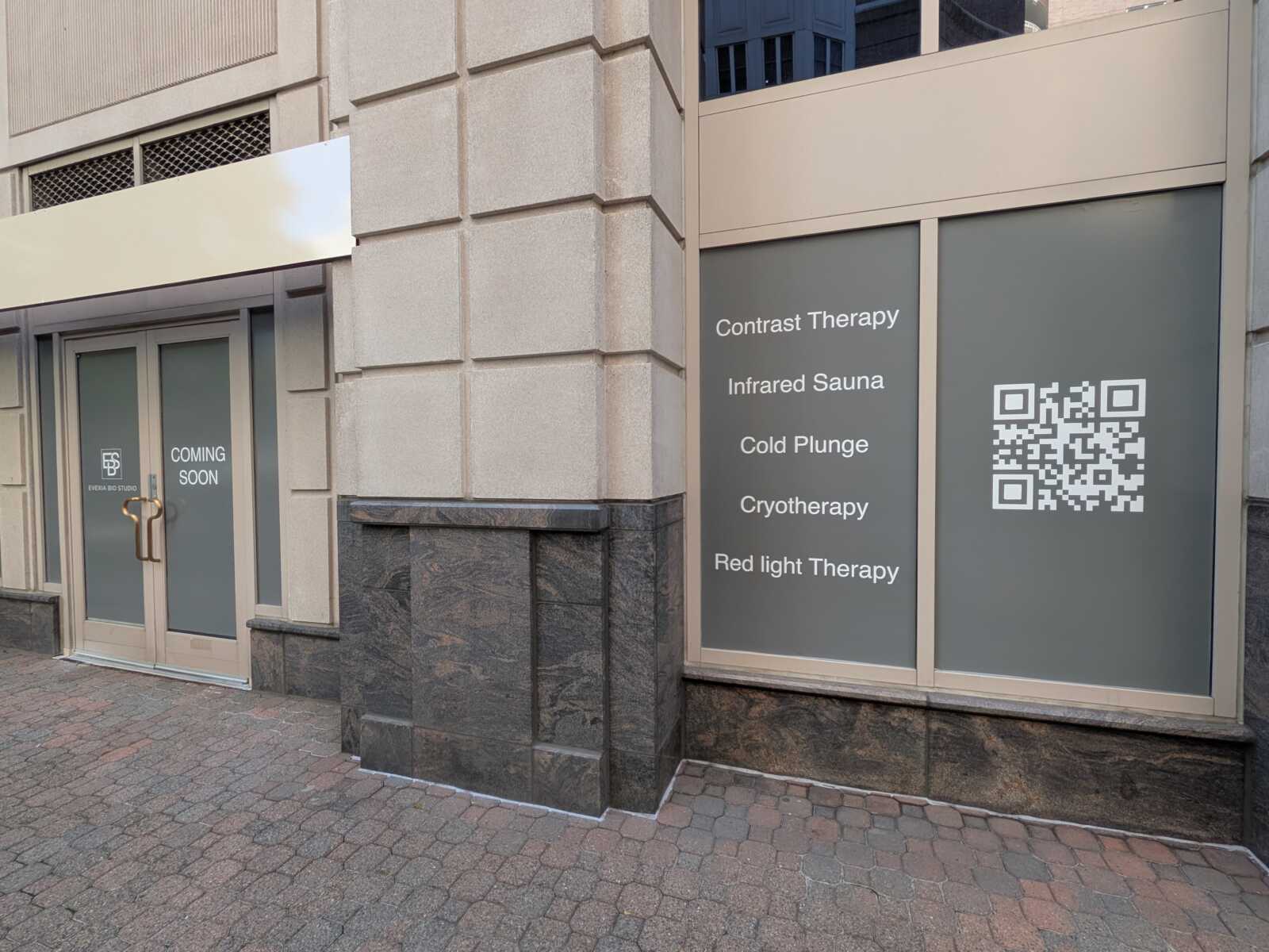 Storefront with a 'Coming Soon' door and a window sign listing wellness services: Contrast Therapy, Infrared Sauna, Cold Plunge, Cryotherapy, Red Light Therapy with a QR code.