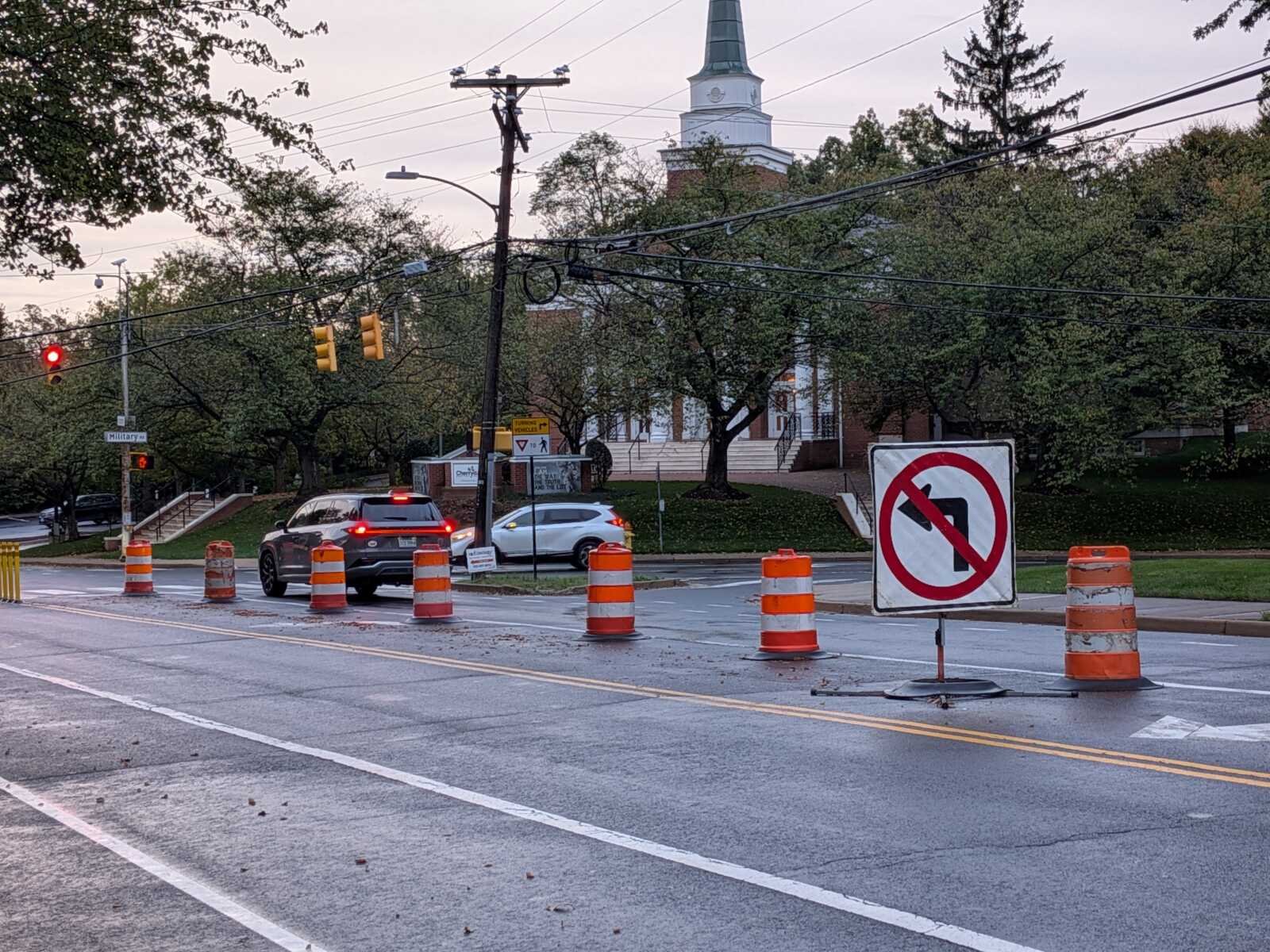 Street under construction with orange traffic barrels blocking lanes; cars drive past with a church steeple in the background.