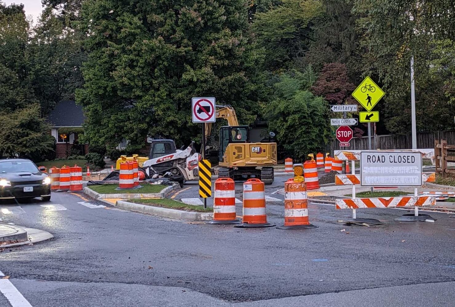 Construction site on a street with orange barrels, stop sign, and 'ROAD CLOSED' sign; heavy equipment in background.