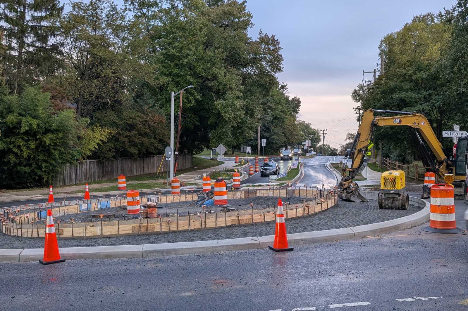 Construction around a roundabout with orange cones, barriers, and a yellow excavator paving the curb area on wet pavement behind a curved curb edge and parked cars farther down the street.