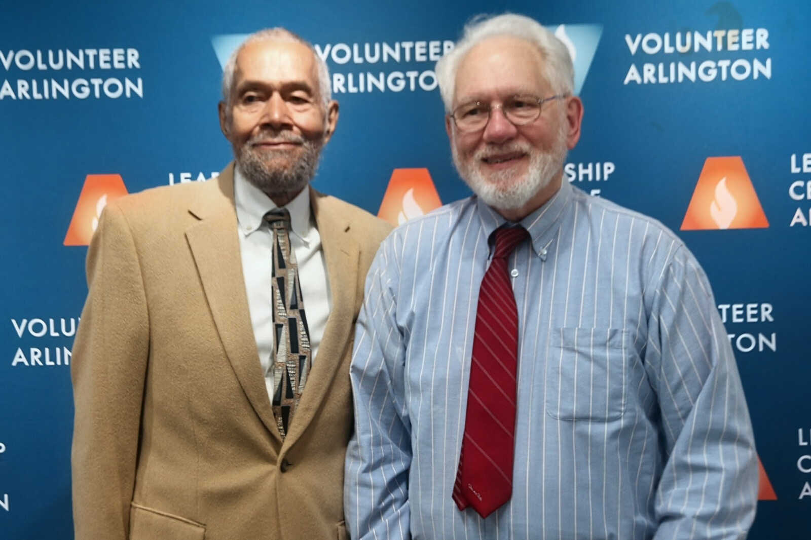 Two older men posing and smiling in front of a blue Volunteer Arlington backdrop; one in a tan suit and patterned tie, the other in a blue striped shirt and red tie.