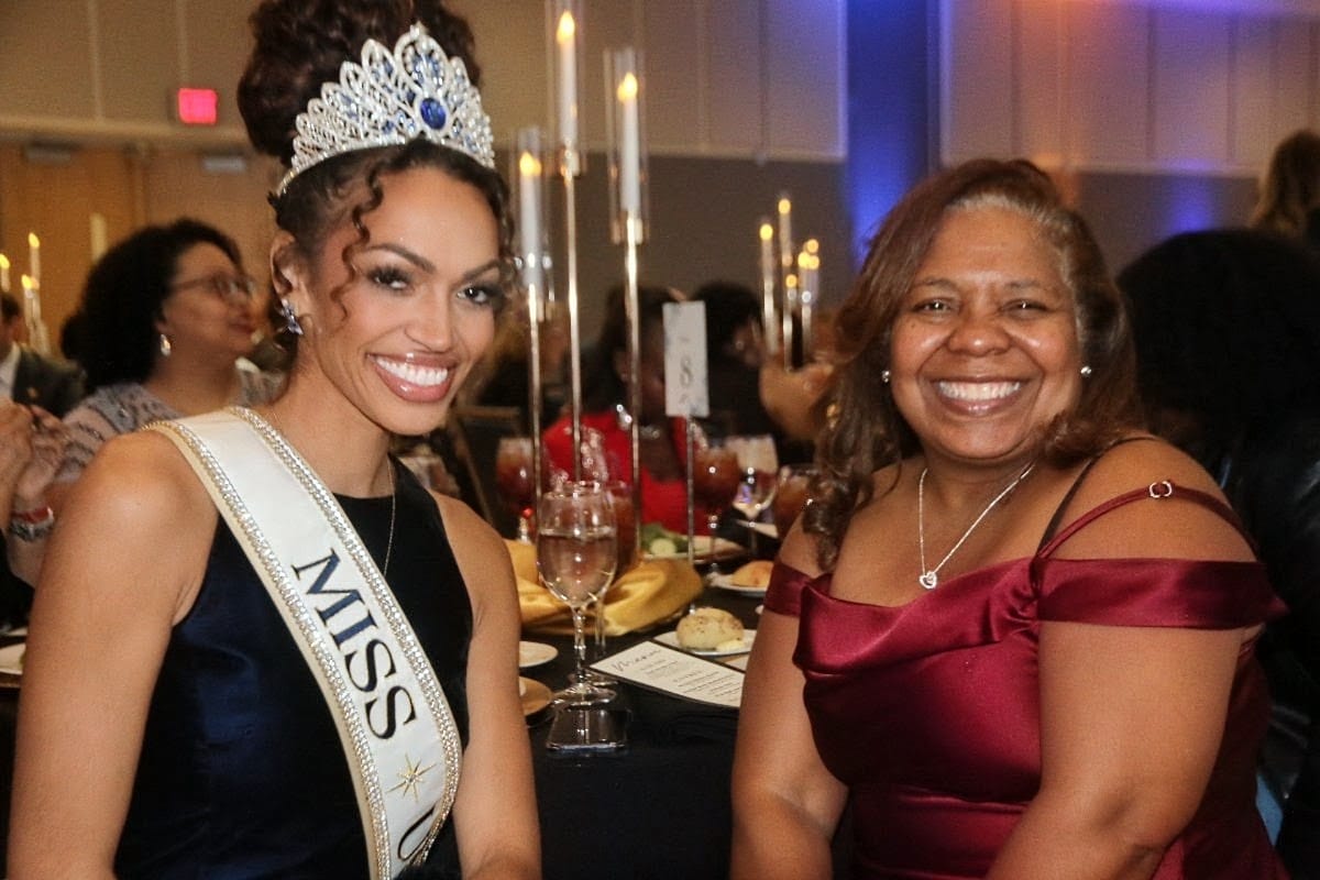 Miss pageant winner wearing a crown and sash sits at a banquet table with a smiling guest at a formal event.