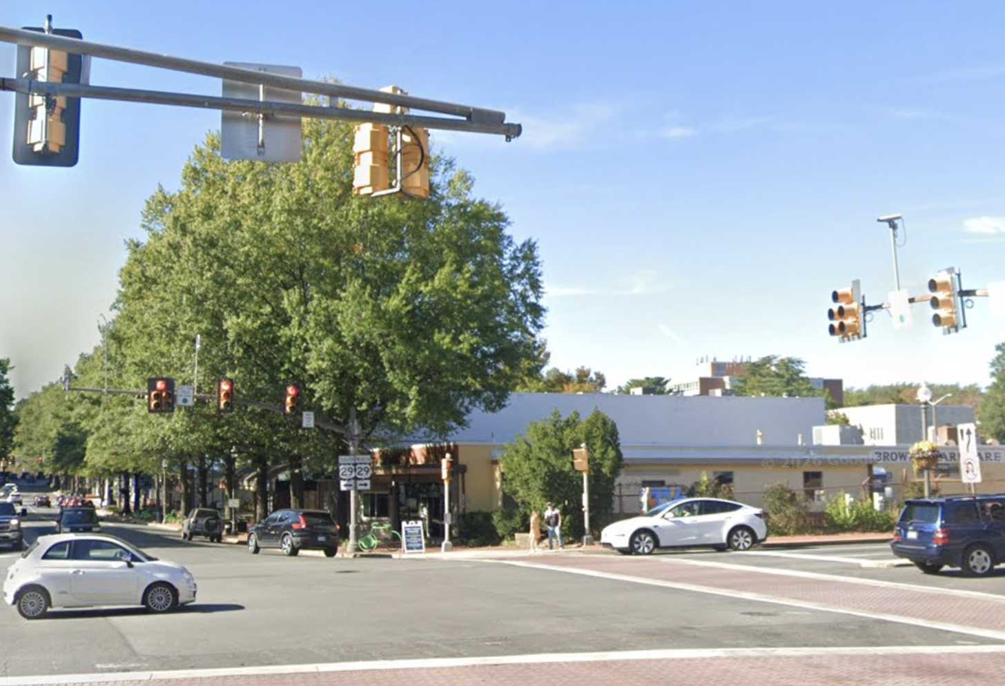 Intersection at a sunny day with traffic lights, cars waiting, and a tree-lined storefront strip across the street.