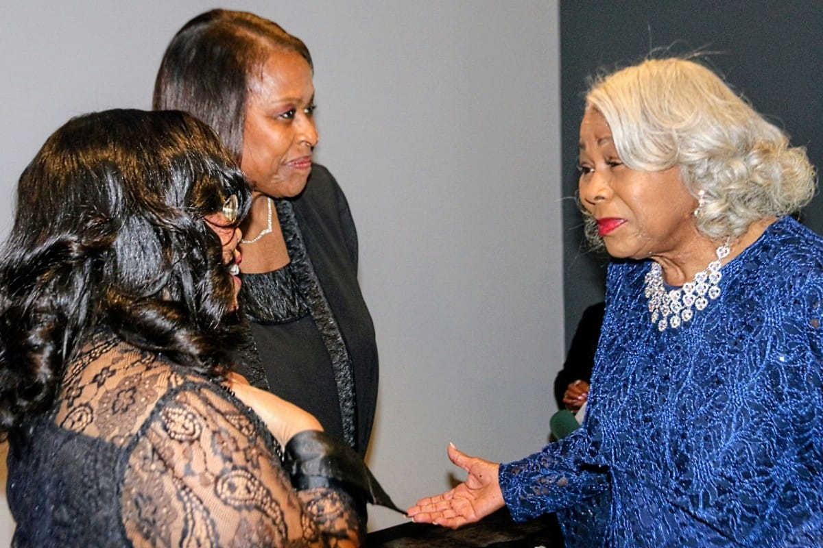 Three women in formal attire greet each other; elderly woman in a blue lace dress extends her hand for a handshake to the others in black outfits.