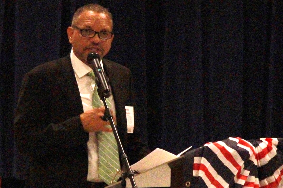 Man in a suit gives a speech at a podium with a microphone, holding papers; a striped flag is draped on the podium.