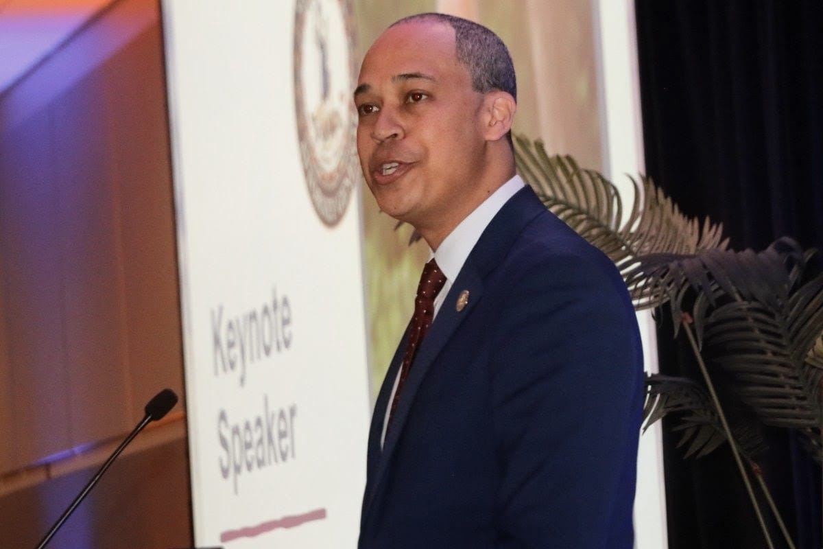 Man in a navy suit delivering a keynote speech on stage, standing at a podium with 'Keynote Speaker' text visible in the background.