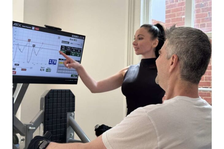 Trainer explains data on a large monitor of a fitness machine to a man seated for a workout assessment.
