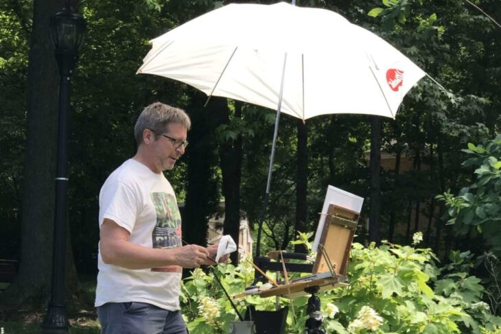 Man painting at an outdoor easel under a white umbrella in a lush garden.