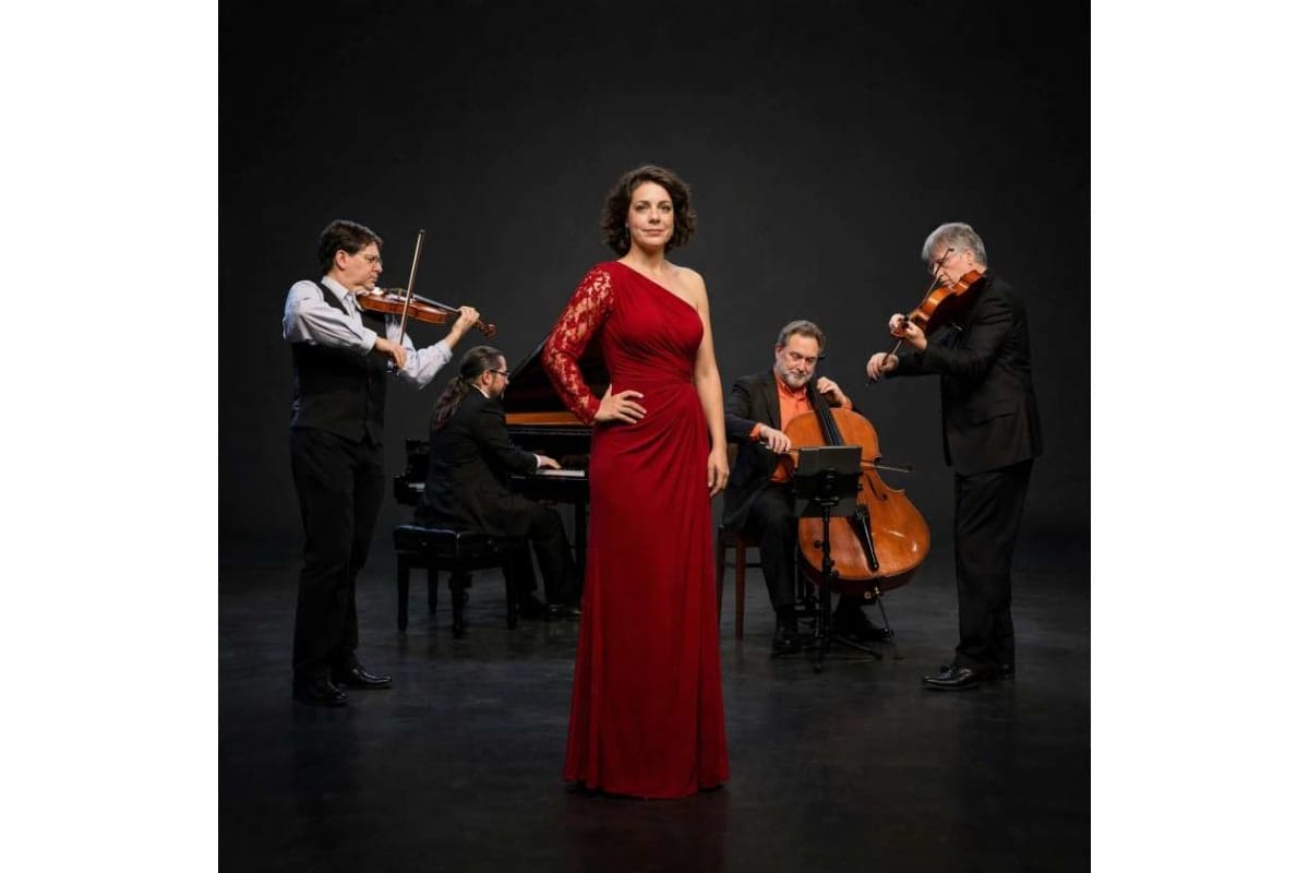 Female singer in a red gown stands center, flanked by a string quartet performing on a dark stage (violinists, pianist, cellist).