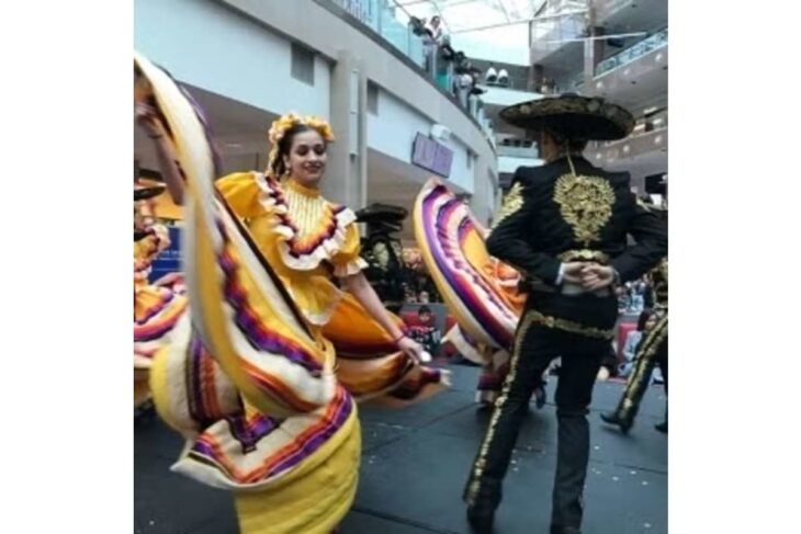 Dancers in colorful folklorico dresses performing a traditional Mexican dance on a stage in a mall.