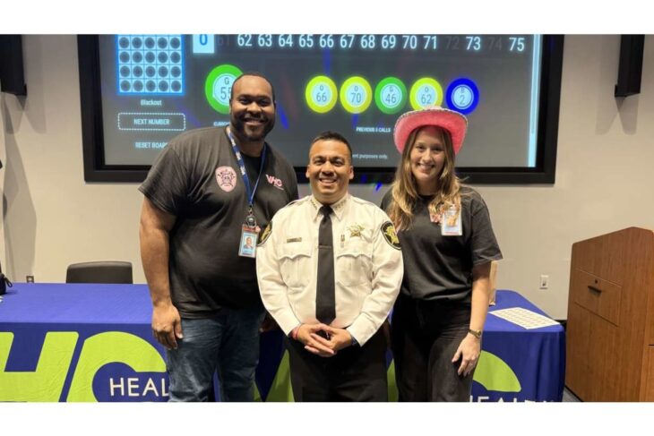 Three people pose at a health booth: a smiling security officer in beige uniform center, flanked by a man in a black shirt and a woman in gray with a pink cowboy hat; a large screen with colorful indicators is behind them.