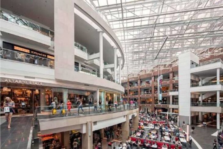 Interior view of a multi-level shopping mall with storefronts, glass railings, and a busy food court below.